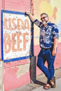 a man in blue jeans, a blue patterned shirt, a beard, and sunglasses, leans casually against a wall where a sign is painted that reads "USDA BEEF".