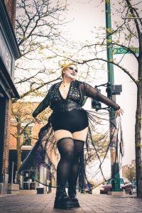 Image of Drag Queen, Baha Blast on a sidewalk, with fall trees behind them, as they are dressed in a black jumpsuit shorts and thigh high, stockings with chunky boots.